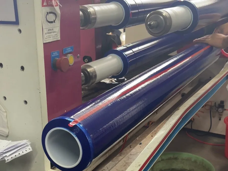 At our factory, a hand smooths a roll of blue protective film on a machine